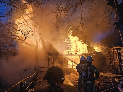 ein brennendes Wochenendhaus mit einem Atemschutztrupp, welcher Löschmaßnahmen einleitet