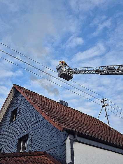 Das Dach eines Hauses mit einem stark qualmenden Schornstein über dem der Drehleiterkorb in Stellung gebracht wird