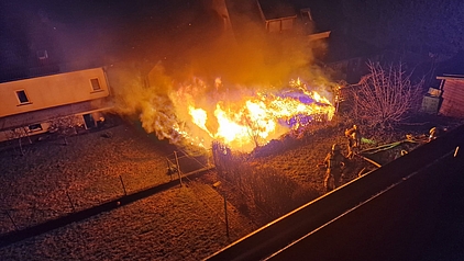 eine brennende Gartenhütte, neben der Feuerwehrleute einen Löschangriff einleiten