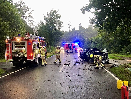 Ein Feuerwehrauto auf einer Landstraße bei einem Verkehrsunfall. Weiterhin zu sehen sind Feuerwehrmänner, ein Krankenwagen und ein verunfalltes, total zerstörtes Auto.