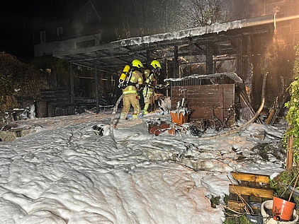 zwei Feuerwehrleute in Schutzausrüstung, die in einem Schaumteppich vor den Überresten einer abgebrannten Gartenhütte stehen