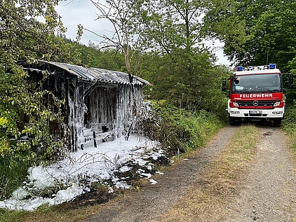 abgelöschter, mit Schaum bedeckter Holzunterstand, neben dem ein Feuerwehrfahrzeug steht. 