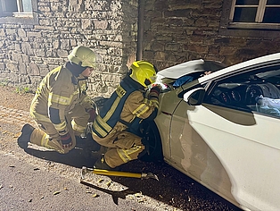 zwei Feuerwehrleute, die an der Motorhaube eines verunfallten PKW knien und hieran arbeiten