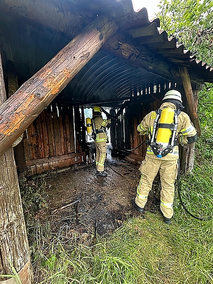 zwei in persönlicher Schutzausrüstung gekleidete Feuerwehrmänner unter Atemschutz beim Löschen eines Holzunterstandes