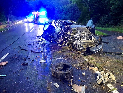 Ein verunfalltes, total zerstörtes Auto auf einer Landstraße. Im Hintergrund steht ein Krankenwagen mit Blaulicht. 